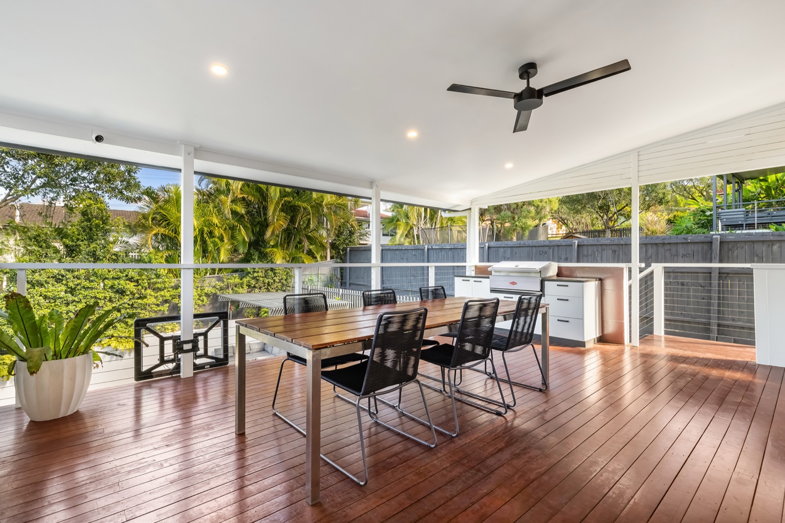 Wide covered deck with a long timber table, ceiling fan and built-in alfresco BBQ kitchen, looking over the leafy backyard