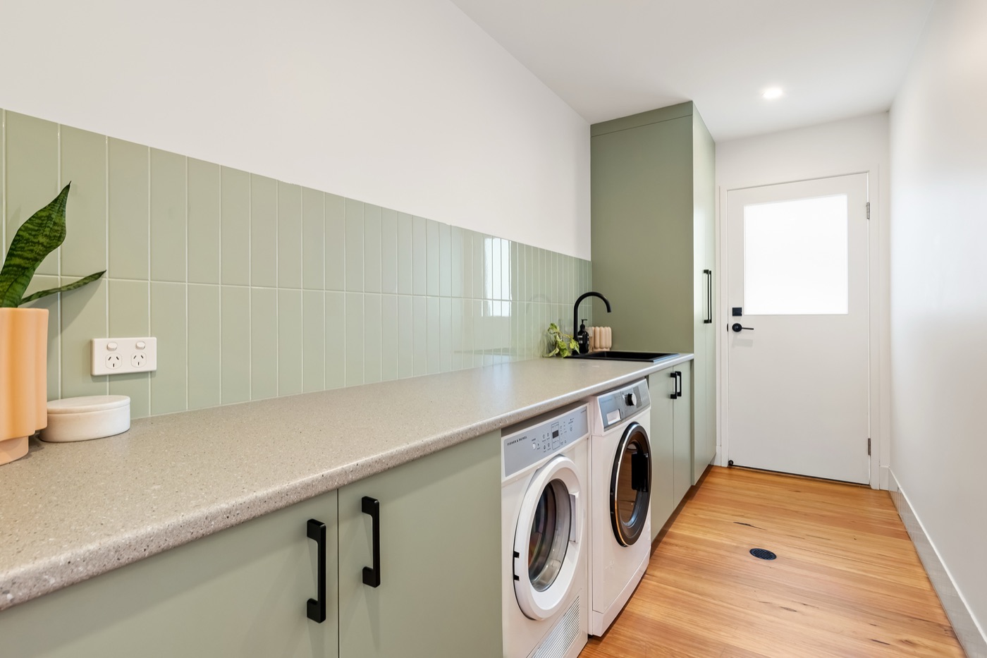 Laundry with sage-green cabinetry, stone benchtop, vertical tiled splashback and side access to the garden