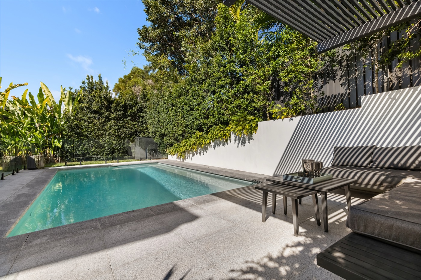 Tiled poolside entertaining area with a daybed under the engineered slat pergola, screened by established gardens
